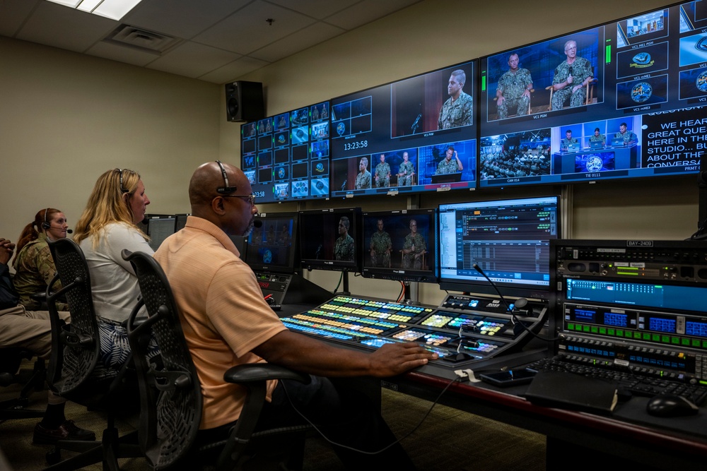 A professional broadcast control room with multiple operators managing a live military production. The room features a large multi-monitor video wall displaying feeds of uniformed Navy personnel, along with a production switcher console and various broadcast equipment. Three visible operators are actively monitoring and controlling the broadcast.