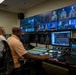 A professional broadcast control room with multiple operators managing a live military production. The room features a large multi-monitor video wall displaying feeds of uniformed Navy personnel, along with a production switcher console and various broadcast equipment. Three visible operators are actively monitoring and controlling the broadcast.