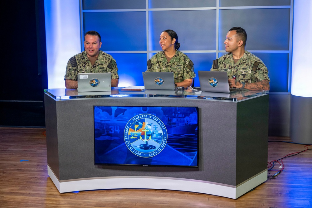 Three U.S. Navy personnel in NWU (Navy Working Uniform) camouflage are seated at a curved broadcast desk in what appears to be a professional television or media studio. Each person has a laptop in front of them displaying a matching logo. A Sharp flat-screen monitor on the front of the desk displays a U.S. Navy emblem reading 'Built in the Foundry · Tempered in the Fleet · Forged to Fight.'