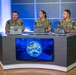 Three U.S. Navy personnel in NWU (Navy Working Uniform) camouflage are seated at a curved broadcast desk in what appears to be a professional television or media studio. Each person has a laptop in front of them displaying a matching logo. A Sharp flat-screen monitor on the front of the desk displays a U.S. Navy emblem reading 'Built in the Foundry · Tempered in the Fleet · Forged to Fight.'