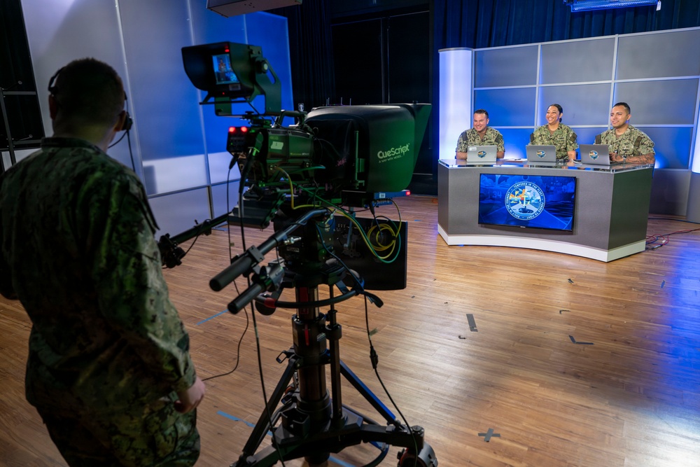 A military television broadcast studio is in operation, with three uniformed personnel seated behind an anchor desk equipped with laptops and a monitor displaying a military emblem. A fourth uniformed individual operates a professional broadcast camera with a teleprompter (CueScript brand) in the foreground. The setting appears to be a professional TV production studio with blue ambient lighting and a modular backdrop.