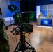 A military television broadcast studio is in operation, with three uniformed personnel seated behind an anchor desk equipped with laptops and a monitor displaying a military emblem. A fourth uniformed individual operates a professional broadcast camera with a teleprompter (CueScript brand) in the foreground. The setting appears to be a professional TV production studio with blue ambient lighting and a modular backdrop.