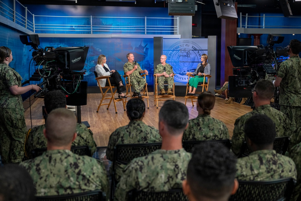 A formal panel discussion or interview is taking place in a professional television studio setting bearing the Department of the Navy seal. Four panelists — two in U.S. Navy uniforms and two civilian women — are seated on director's chairs on a broadcast stage. An audience of Navy personnel in camouflage uniforms observes from the foreground while camera operators record the event.