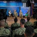 A formal panel discussion or interview is taking place in a professional television studio setting bearing the Department of the Navy seal. Four panelists — two in U.S. Navy uniforms and two civilian women — are seated on director's chairs on a broadcast stage. An audience of Navy personnel in camouflage uniforms observes from the foreground while camera operators record the event.