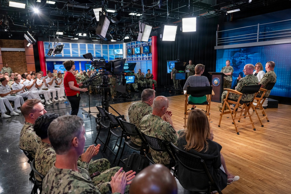 A military event or broadcast taping is taking place in a professional television studio setting. A large audience of uniformed U.S. Navy personnel and some civilians are seated and applauding, while a panel of individuals — including military officers and civilians — are seated on a stage area. A woman in a red top appears to be hosting or moderating the event near a professional broadcast camera.
