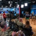 A military event or broadcast taping is taking place in a professional television studio setting. A large audience of uniformed U.S. Navy personnel and some civilians are seated and applauding, while a panel of individuals — including military officers and civilians — are seated on a stage area. A woman in a red top appears to be hosting or moderating the event near a professional broadcast camera.