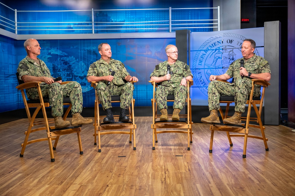 Four U.S. Navy personnel in NWU Type III (woodland camouflage) uniforms are seated on wooden folding chairs in what appears to be a professional broadcast studio. The setting includes a backdrop displaying the U.S. Department of the Navy seal and a blue digital display background. The individuals appear to be participating in a panel discussion or recorded interview, with at least two holding microphones.