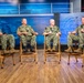 Four U.S. Navy personnel in NWU Type III (woodland camouflage) uniforms are seated on wooden folding chairs in what appears to be a professional broadcast studio. The setting includes a backdrop displaying the U.S. Department of the Navy seal and a blue digital display background. The individuals appear to be participating in a panel discussion or recorded interview, with at least two holding microphones.
