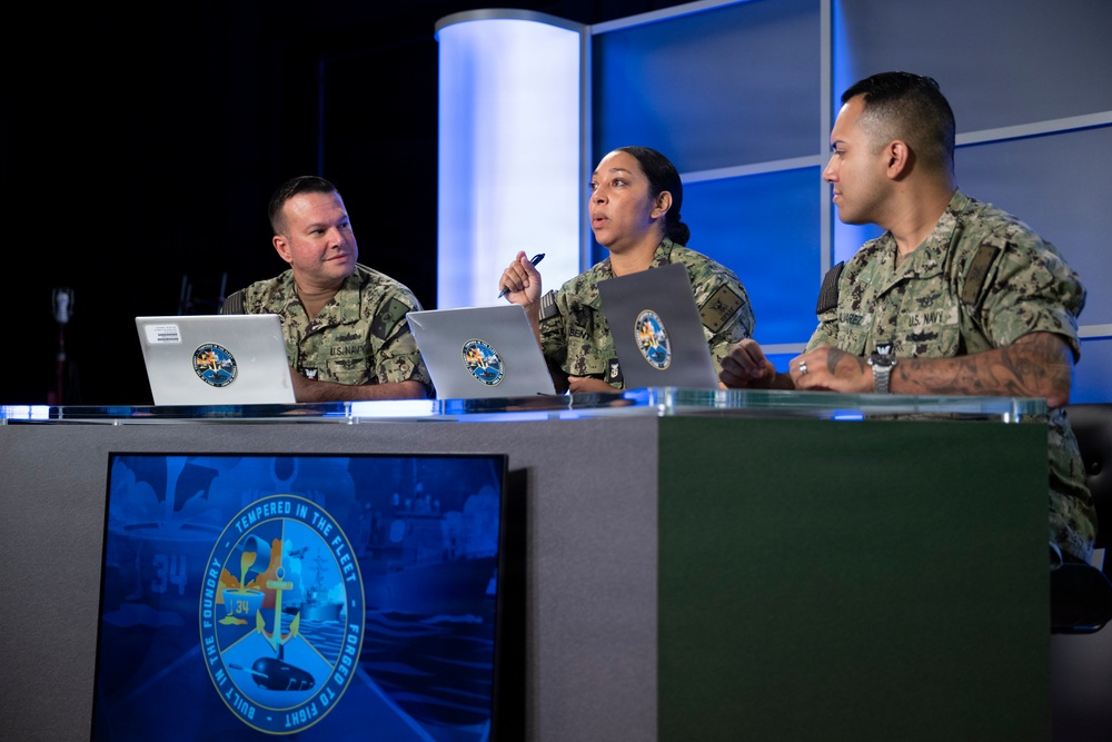 Three U.S. Navy personnel in NWU (Navy Working Uniform) camouflage are seated at a studio desk with laptops, appearing to participate in a broadcast or panel discussion. The central figure, a female sailor, is speaking and holding a pen, while the other two listen. A display screen in the foreground shows an emblem reading 'Built in the Foundry – Tempered in the Fleet – Forged to Fight' with the number 34.
