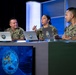 Three U.S. Navy personnel in NWU (Navy Working Uniform) camouflage are seated at a studio desk with laptops, appearing to participate in a broadcast or panel discussion. The central figure, a female sailor, is speaking and holding a pen, while the other two listen. A display screen in the foreground shows an emblem reading 'Built in the Foundry – Tempered in the Fleet – Forged to Fight' with the number 34.