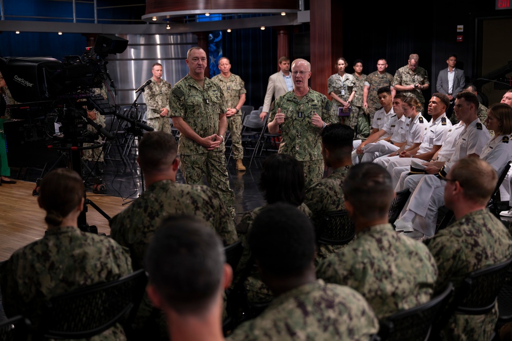 Two senior U.S. Navy officers in camouflage NWU (Navy Working Uniform) are addressing a mixed audience of enlisted sailors and naval officers in white dress uniforms. The setting appears to be a media or briefing studio, indicated by professional broadcast cameras and teleprompter equipment visible on the left. The gathering resembles an all-hands meeting, town hall, or leadership engagement session.