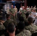 Two senior U.S. Navy officers in camouflage NWU (Navy Working Uniform) are addressing a mixed audience of enlisted sailors and naval officers in white dress uniforms. The setting appears to be a media or briefing studio, indicated by professional broadcast cameras and teleprompter equipment visible on the left. The gathering resembles an all-hands meeting, town hall, or leadership engagement session.