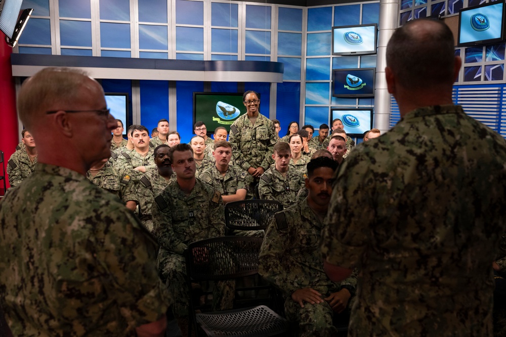 A group of U.S. Navy personnel in NWU (Navy Working Uniform) camouflage are gathered in what appears to be a Navy media or broadcast studio. Two senior personnel, visible from behind in the foreground, appear to be addressing the assembled sailors. The setting features multiple monitors displaying the U.S. Navy seal and a professional broadcast studio backdrop.