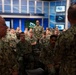 A group of U.S. Navy personnel in NWU (Navy Working Uniform) camouflage are gathered in what appears to be a Navy media or broadcast studio. Two senior personnel, visible from behind in the foreground, appear to be addressing the assembled sailors. The setting features multiple monitors displaying the U.S. Navy seal and a professional broadcast studio backdrop.