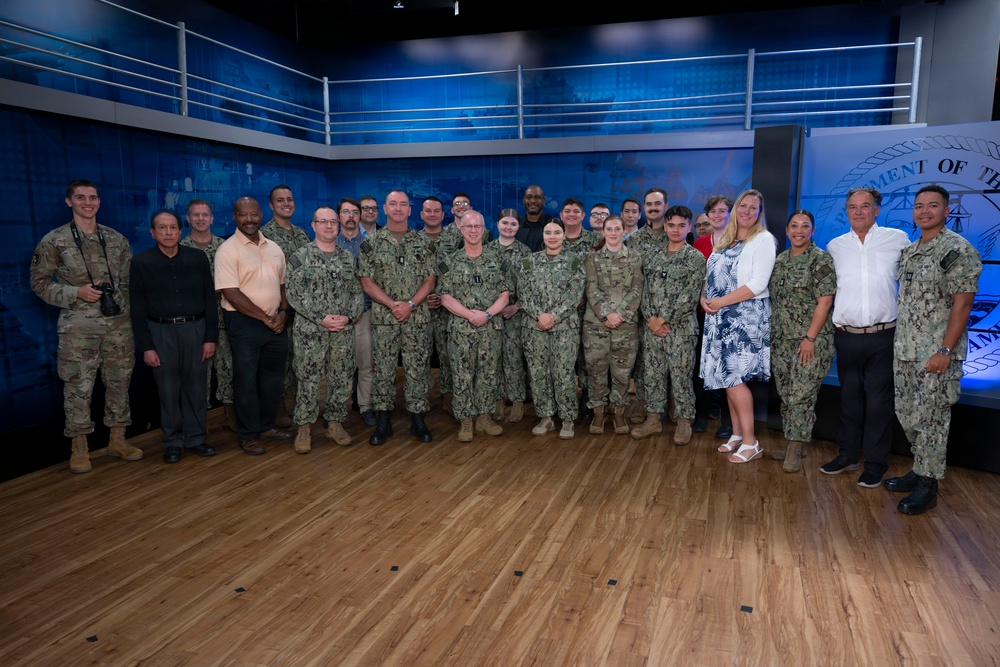 A large group of approximately 30 individuals poses for a formal group photograph in what appears to be a military media studio or briefing room. The group consists primarily of U.S. Navy personnel in digital camouflage uniforms, along with a few civilians and one Army soldier. A partial Department of the Navy seal is visible on the right side of the background.