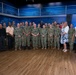 A large group of approximately 30 individuals poses for a formal group photograph in what appears to be a military media studio or briefing room. The group consists primarily of U.S. Navy personnel in digital camouflage uniforms, along with a few civilians and one Army soldier. A partial Department of the Navy seal is visible on the right side of the background.