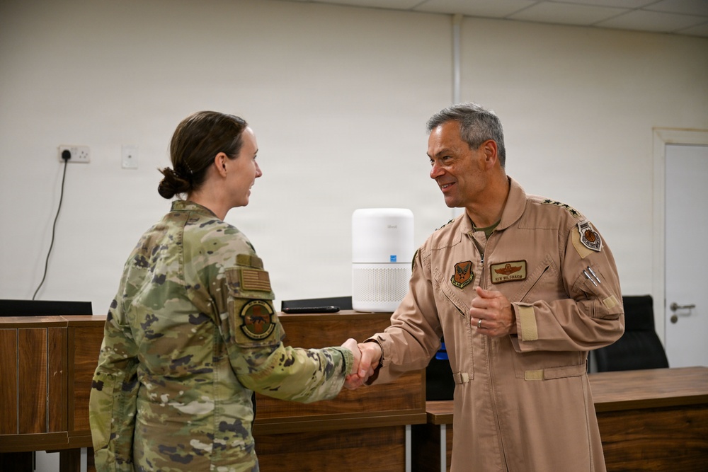Two military personnel are shaking hands in what appears to be an indoor briefing or conference room setting. The individual on the left is wearing a multicam/OCP camouflage uniform, while the individual on the right is wearing a tan/desert flight suit with visible patches and rank insignia. The interaction appears formal and ceremonial in nature.