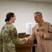 Two military personnel are shaking hands in what appears to be an indoor briefing or conference room setting. The individual on the left is wearing a multicam/OCP camouflage uniform, while the individual on the right is wearing a tan/desert flight suit with visible patches and rank insignia. The interaction appears formal and ceremonial in nature.