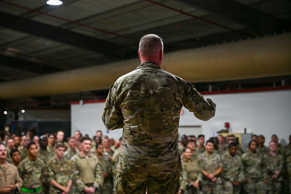 A military officer or senior NCO is addressing a large formation of uniformed personnel inside what appears to be a hangar or large industrial facility. The speaker is photographed from behind, facing a crowd of dozens of soldiers or airmen all wearing matching camouflage uniforms. The setting and formation suggest an official briefing, commander's call, or unit assembly.