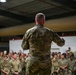 A military officer or senior NCO is addressing a large formation of uniformed personnel inside what appears to be a hangar or large industrial facility. The speaker is photographed from behind, facing a crowd of dozens of soldiers or airmen all wearing matching camouflage uniforms. The setting and formation suggest an official briefing, commander's call, or unit assembly.