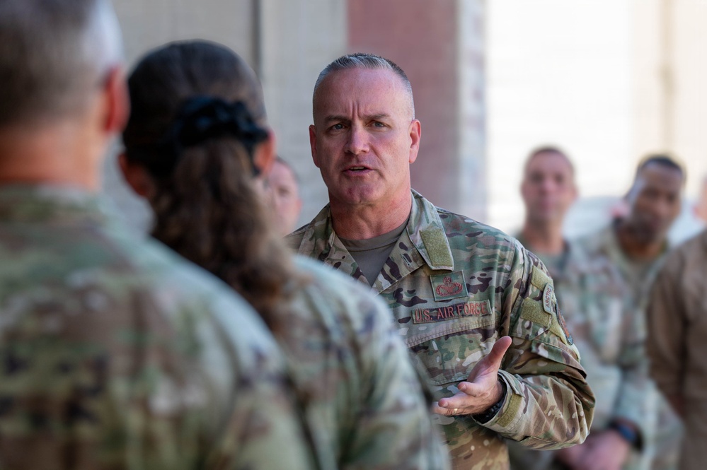 A U.S. Air Force service member in Operational Camouflage Pattern (OCP) uniform is addressing a group of fellow military personnel outdoors. The central figure appears to be speaking or briefing the group, gesturing with one hand in what appears to be an informal gathering or formation. Multiple uniformed personnel are visible in the background observing the exchange.