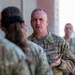 A U.S. Air Force service member in Operational Camouflage Pattern (OCP) uniform is addressing a group of fellow military personnel outdoors. The central figure appears to be speaking or briefing the group, gesturing with one hand in what appears to be an informal gathering or formation. Multiple uniformed personnel are visible in the background observing the exchange.