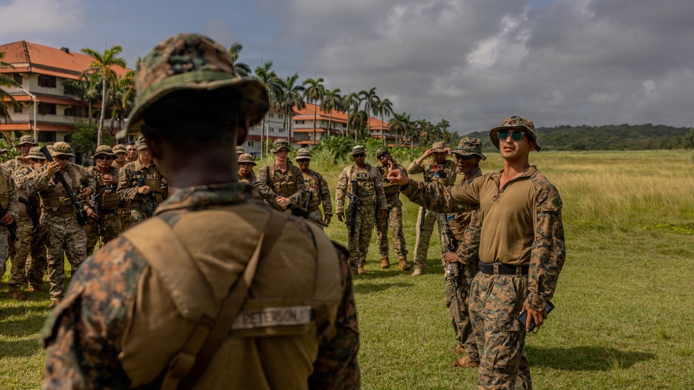 A group of military personnel in camouflage uniforms are assembled on a grassy field, apparently receiving a briefing or instruction from a senior figure wearing sunglasses and a tan shirt. The setting appears to be a tropical or subtropical location, evidenced by palm trees and red-roofed buildings in the background. The foreground shows a soldier with the name tape 'PETERSON' visible on their uniform.