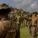 A group of military personnel in camouflage uniforms are assembled on a grassy field, apparently receiving a briefing or instruction from a senior figure wearing sunglasses and a tan shirt. The setting appears to be a tropical or subtropical location, evidenced by palm trees and red-roofed buildings in the background. The foreground shows a soldier with the name tape 'PETERSON' visible on their uniform.
