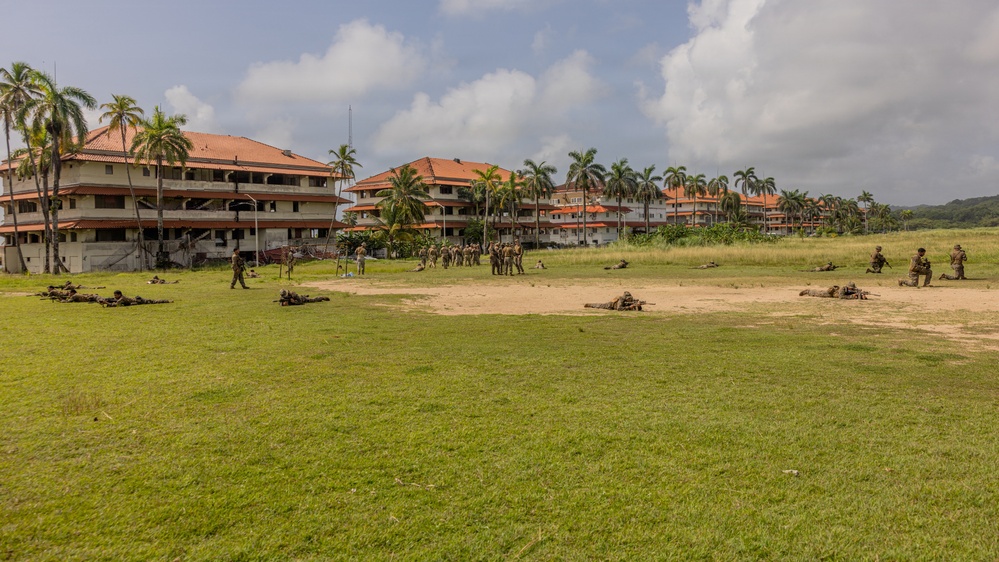 Military personnel are conducting a field training exercise on a large grassy area adjacent to multi-story buildings with terracotta roofs, surrounded by palm trees in a tropical setting. Soldiers are dispersed across the field in various tactical positions, including prone firing positions and standing formations. The environment suggests a military installation in a tropical or subtropical region.