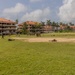Military personnel are conducting a field training exercise on a large grassy area adjacent to multi-story buildings with terracotta roofs, surrounded by palm trees in a tropical setting. Soldiers are dispersed across the field in various tactical positions, including prone firing positions and standing formations. The environment suggests a military installation in a tropical or subtropical region.
