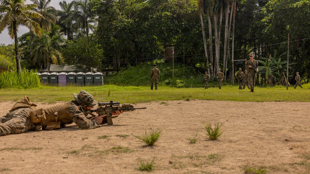 Military personnel are conducting what appears to be a live-fire range exercise in a tropical environment. One soldier is in a prone shooting position in the foreground aiming a scoped rifle downrange, while several other personnel are visible in the background moving across the field. The setting includes portable toilets, a flagpole, and dense tropical vegetation consistent with a training base in a tropical region.