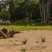 Military personnel are conducting what appears to be a live-fire range exercise in a tropical environment. One soldier is in a prone shooting position in the foreground aiming a scoped rifle downrange, while several other personnel are visible in the background moving across the field. The setting includes portable toilets, a flagpole, and dense tropical vegetation consistent with a training base in a tropical region.