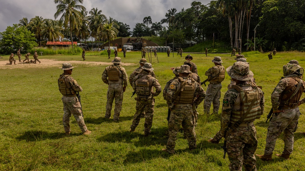 A group of military personnel in camouflage uniforms and tactical gear are gathered on a grassy field in a tropical environment, appearing to observe or receive a briefing. The setting includes palm trees, jungle vegetation, and what appears to be a forward operating base or training facility in the background. The scene suggests a multinational military training exercise or field operation.