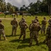 A group of military personnel in camouflage uniforms and tactical gear are gathered on a grassy field in a tropical environment, appearing to observe or receive a briefing. The setting includes palm trees, jungle vegetation, and what appears to be a forward operating base or training facility in the background. The scene suggests a multinational military training exercise or field operation.