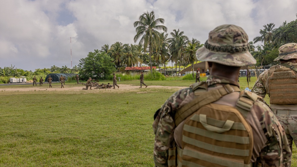 Military personnel are conducting field training exercises in a tropical environment. Two soldiers in the foreground wearing multicam uniforms and tactical gear observe a group of soldiers in the background performing drills on a sandy training area. The setting includes palm trees, temporary structures, and what appears to be a forward operating base or training compound.