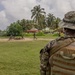 Military personnel are conducting field training exercises in a tropical environment. Two soldiers in the foreground wearing multicam uniforms and tactical gear observe a group of soldiers in the background performing drills on a sandy training area. The setting includes palm trees, temporary structures, and what appears to be a forward operating base or training compound.