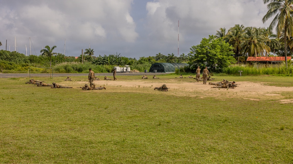 Military personnel in camouflage uniforms are conducting a field training exercise on an open grassy area with a sandy patch. Multiple soldiers are in prone positions while a few are standing, suggesting a tactical drill or marksmanship training. The setting appears to be a tropical military installation based on the surrounding vegetation and infrastructure.