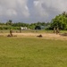 Military personnel in camouflage uniforms are conducting a field training exercise on an open grassy area with a sandy patch. Multiple soldiers are in prone positions while a few are standing, suggesting a tactical drill or marksmanship training. The setting appears to be a tropical military installation based on the surrounding vegetation and infrastructure.