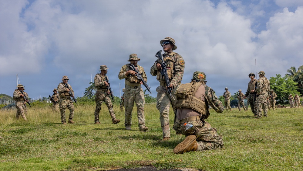 A group of military personnel in multicam/OCP camouflage uniforms are conducting what appears to be a field training exercise on a grassy open area in a tropical environment. Soldiers are equipped with rifles, tactical vests, and boonie hats, with one individual kneeling in the foreground while others stand and move in the background. The setting suggests a warm-weather military installation, likely in a Pacific or tropical region based on the vegetation.