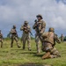 A group of military personnel in multicam/OCP camouflage uniforms are conducting what appears to be a field training exercise on a grassy open area in a tropical environment. Soldiers are equipped with rifles, tactical vests, and boonie hats, with one individual kneeling in the foreground while others stand and move in the background. The setting suggests a warm-weather military installation, likely in a Pacific or tropical region based on the vegetation.