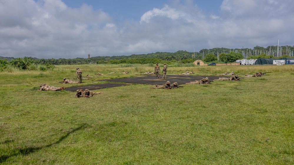 Military personnel in camouflage uniforms are conducting a prone shooting or tactical training exercise on and around a dark paved surface in an open grassy field. Several soldiers are lying in the prone position while two or three individuals appear to be standing as instructors or supervisors. The setting includes a tropical or subtropical environment with dense tree lines, military tents, and support vehicles visible in the background.