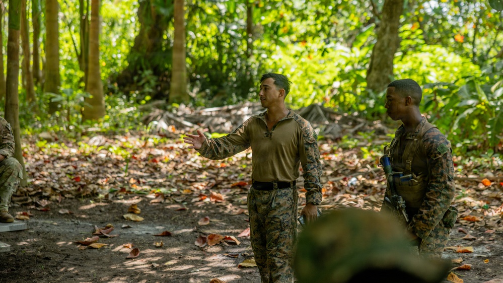 Two military personnel in camouflage uniforms are engaged in what appears to be a briefing or instruction session in a jungle or forested training environment. One individual is gesturing with an outstretched hand, apparently explaining or directing, while the other listens attentively. Additional personnel are partially visible at the edges of the frame.