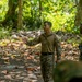 Two military personnel in camouflage uniforms are engaged in what appears to be a briefing or instruction session in a jungle or forested training environment. One individual is gesturing with an outstretched hand, apparently explaining or directing, while the other listens attentively. Additional personnel are partially visible at the edges of the frame.
