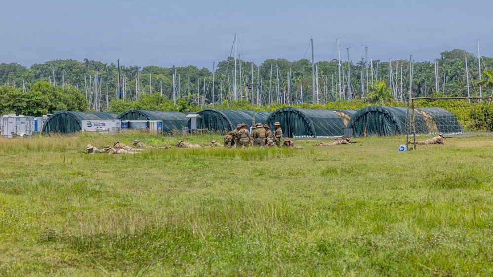 A group of military personnel in camouflage uniforms are conducting what appears to be a field training exercise on an open grassy area. Several soldiers are prone on the ground while others stand in a small cluster near a row of large green military tent structures. The background features dense tropical vegetation, tall masts (likely from a marina), and utility poles.