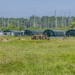 A group of military personnel in camouflage uniforms are conducting what appears to be a field training exercise on an open grassy area. Several soldiers are prone on the ground while others stand in a small cluster near a row of large green military tent structures. The background features dense tropical vegetation, tall masts (likely from a marina), and utility poles.