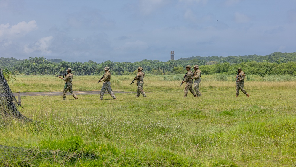 Five armed military personnel in camouflage uniforms are walking in a loose patrol formation across an open grassy field. The setting appears to be a tropical or subtropical environment, indicated by the dense palm and broadleaf tree line in the background. A tall concrete or stone tower structure is visible in the distance among the trees.