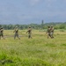 Five armed military personnel in camouflage uniforms are walking in a loose patrol formation across an open grassy field. The setting appears to be a tropical or subtropical environment, indicated by the dense palm and broadleaf tree line in the background. A tall concrete or stone tower structure is visible in the distance among the trees.