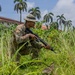 A soldier in camouflage uniform and boonie hat is crouching in tall grass while holding a rifle in a low-ready position. The setting appears to be a tropical or subtropical environment, indicated by palm trees, dense vegetation, and a building with a red roof in the background. The scene suggests a military training exercise or patrol operation.