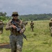 A group of military personnel in camouflage uniforms are conducting a patrol or field exercise across an open grassy area in what appears to be a tropical environment. The foreground soldier is prominently carrying an assault rifle and wearing tactical gear including a plate carrier vest and boonie hat. The setting features lush vegetation, palm trees, and overcast skies consistent with a tropical or subtropical location.