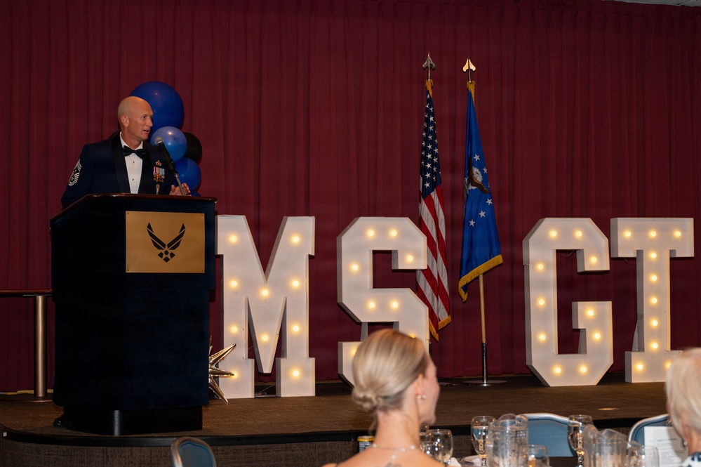 A senior U.S. Air Force enlisted member, identifiable by Chief Master Sergeant rank insignia on his mess dress uniform, delivers a speech at a formal military banquet. The event appears to be an MSGT (Master Sergeant) promotion ceremony or recognition dinner, indicated by illuminated marquee letters spelling 'MSGT' on stage. The setting includes an audience seated at formal dining tables, blue and black balloons, and both the U.S. flag and Air Force flag displayed on stage.